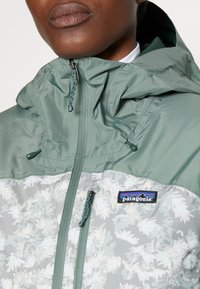 Zelená bunda má kapuci, dolní panel s květinovým vzorem a přední zip. Na hrudi obsahuje logo Patagonia.