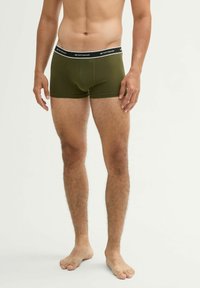 Mannelijke model draagt olijfgroene boxershorts met een zwarte tailleband met witte Tom Tailor branding, staande op blote voeten tegen een witte achtergrond.