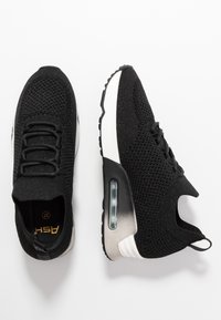 Chaussures de sport en tricot noir avec semelles blanches amorties à air, une chaussure vue de dessus et l'autre vue de côté sur un fond blanc.