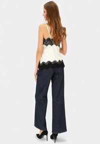 Kremowa koszulka na ramiączkach z koronkowym wykończeniem i czarnymi koronkowymi akcentami, zestawiona z szerokimi ciemnymi dżinsowymi spodniami. Modelka ma na sobie czarne szpilki.
