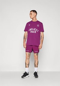 Hombre con tatuajes que lleva una camiseta morada de "Project Rock" y pantalones cortos a juego, calcetines blancos y zapatillas negras, de pie contra un fondo liso.