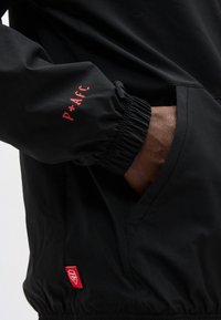Manica di giacca nera con scritta rossa "P+AFC", mano nella tasca laterale e piccolo logo rosso dell'Arsenal vicino all'orlo elastico.