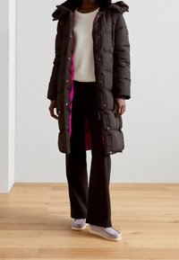 Černá péřová bunda s růžovou podšívkou, s kapucí a patentkami, nošená přes krémový svetr a černé kalhoty, doplněná o světlé boty.