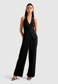 Woman with long dark hair wears sleeveless black halter top and wide-leg black pants with black high-heeled sandals on light gray background.