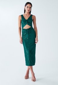 Smaragdinės žalios spalvos blizganti suknelė su aptemptu kirpimu, V formos iškirpte, išpjova liemens srityje ir raišteliais juosmens zonoje. Lygi, blizgi tekstūra.