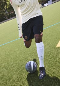 Träningsutrustning som består av en krämfärgad långärmad topp, svarta shorts med en logotyp, vita strumpor och svarta fotbollsskor på ett grönt konstgräsfält.