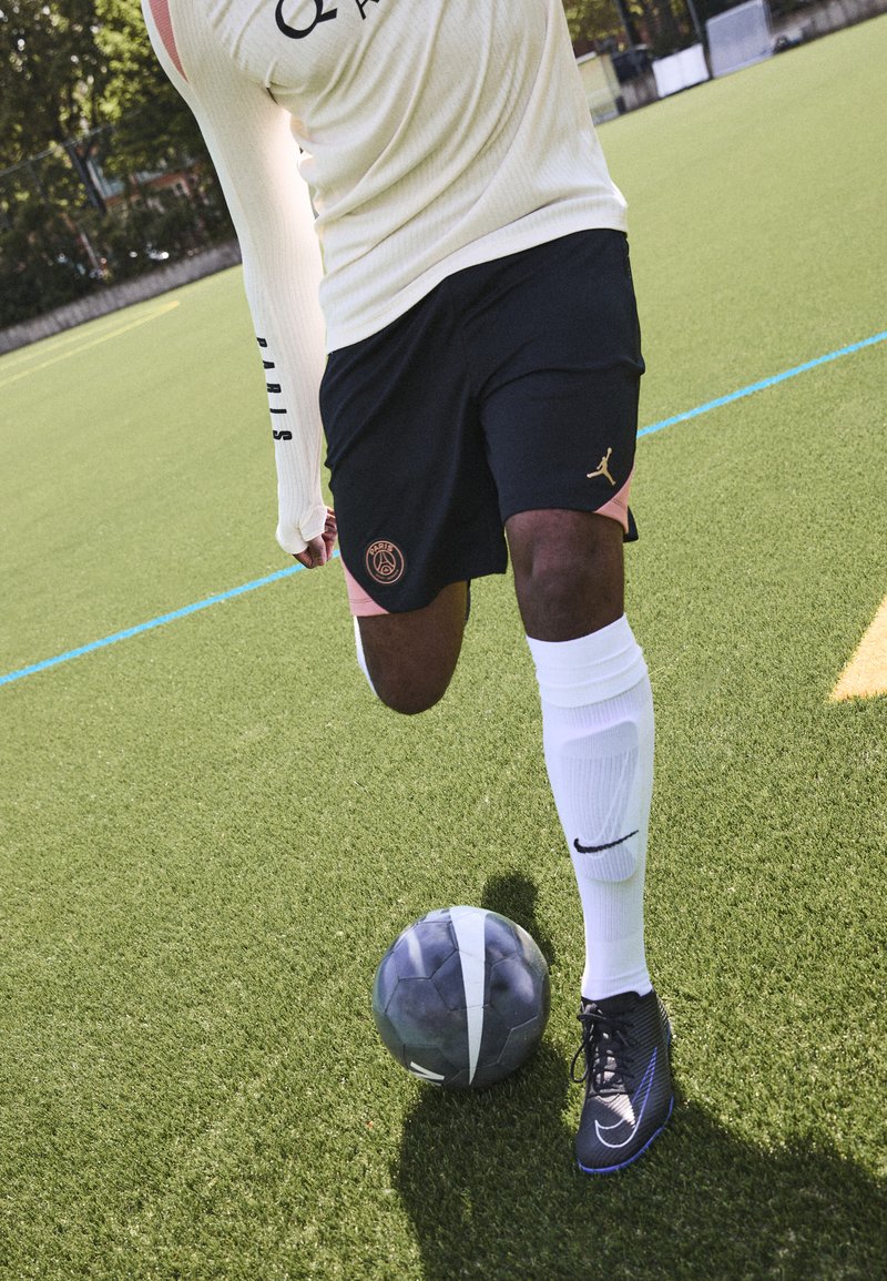 Träningsutrustning som består av en krämfärgad långärmad topp, svarta shorts med en logotyp, vita strumpor och svarta fotbollsskor på ett grönt konstgräsfält.