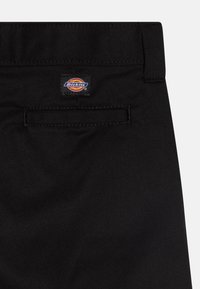 Črne delovne hlače z pravokotno zadnjo žepom in pisano nalepko Dickies na pasu. Tkanina se zdi trpežna in teksturirana.