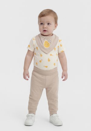 Småbarn i beige bukser, hvid skjorte med gule pæreprint, en beige hagesmæk med en smilende pære og hvide sneakers, der står på en hvid baggrund.