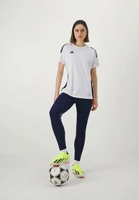 Bela športna majica z črnimi detajli, mornarsko modre hlače in svetlo rumeni nogometni čevlji. Črno-bela nogometna žoga je postavljena ob nogah.