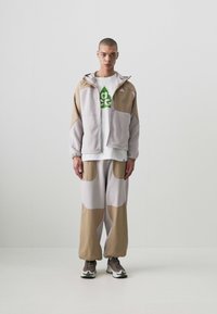 Lys grå og tan fleece treningstøy med overdimensjonert jakke og løse bukser. Grønt grafisk design på hvit t-skjorte, parret med nøytrale atletiske sko.