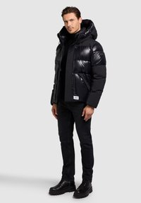 Čierna vatovaná bunda s lesklým povrchom, predĺžená kapucňa, predný zips a bočné vrecká. Nosená s čiernymi džínsami a čiernymi šnurovacími čižmami.