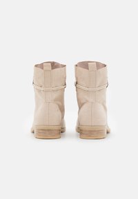 Botas de tobillo de ante beige con suelas de madera, vistas desde atrás sobre un fondo blanco, con tiradores y correas alrededor.