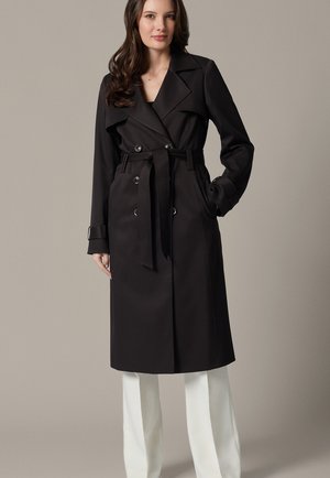 Kobieta ubrana w długi czarny trenčkot z paskiem na białych spodniach, stojąca z jedną ręką w kieszeni na jednolitym neutralnym tle.