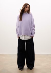 Lavanda oversized pulover s okruglim izrezom, preko bijele košulje, u kombinaciji s širokim tamnoplavi trapericama i crnim cipelama s špicastim vrhom.