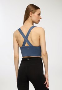 Sport bra blu con spalline incrociate, realizzato in tessuto elasticizzato. Texture liscia, branding minimale, indossato con leggings neri.