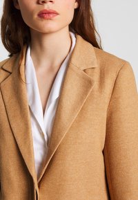 Blazer de textura beige con diseño entallado. Presenta solapas con muesca, un cierre de un solo botón y una camisa blanca con cuello debajo.