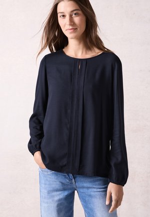 Marineblauwe blouse met lange mouwen, gemaakt van gestructureerde stof, ronde hals en centrale plooidetail, gecombineerd met lichtblauwe jeans.