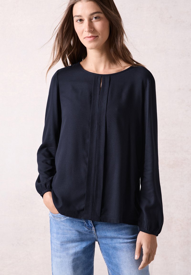 Blusa a maniche lunghe blu navy in tessuto testurizzato, con scollatura rotonda e dettaglio centrale in piega, abbinata a jeans azzurri.