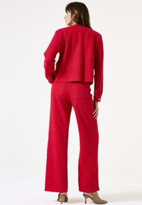 Un costume bicolore rouge texturé comprenant une veste courte avec des manches longues et un pantalon à jambes larges avec deux poches arrière.