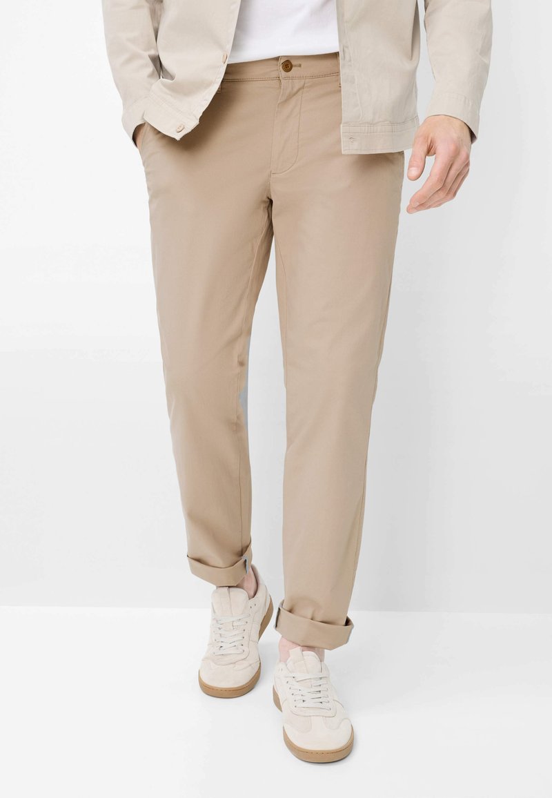 Man draagt beige omslagbroeken, witte sneakers en een lichtbeige jas, met handen deels zichtbaar aan de zijkanten.