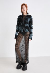 Jachetă neagră și teal cu dungi, cu fermoar, purtată peste o rochie tricotată neagră transparentă cu un model. Pantofi cu platformă masivă, negri, completează ținuta.
