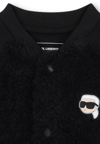 Giubbotto bomber nero con texture pelosa, colletto a coste e bottoni frontali. Presenta un logo a cartone animato di una figura con occhiali da sole sul petto.