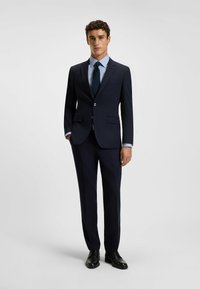 Completo blu con un blazer strutturato, rever a punta e due tasche frontali, abbinato a una camicia a righe blu chiaro e una cravatta scura, completato da scarpe nere.