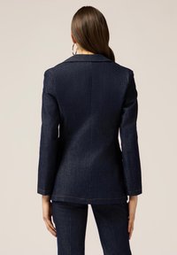 Blazer in denim scuro con una vestibilità strutturata, colletto a bavero e chiusura con un solo bottone. Presenta cuciture visibili e una silhouette sartoriale.