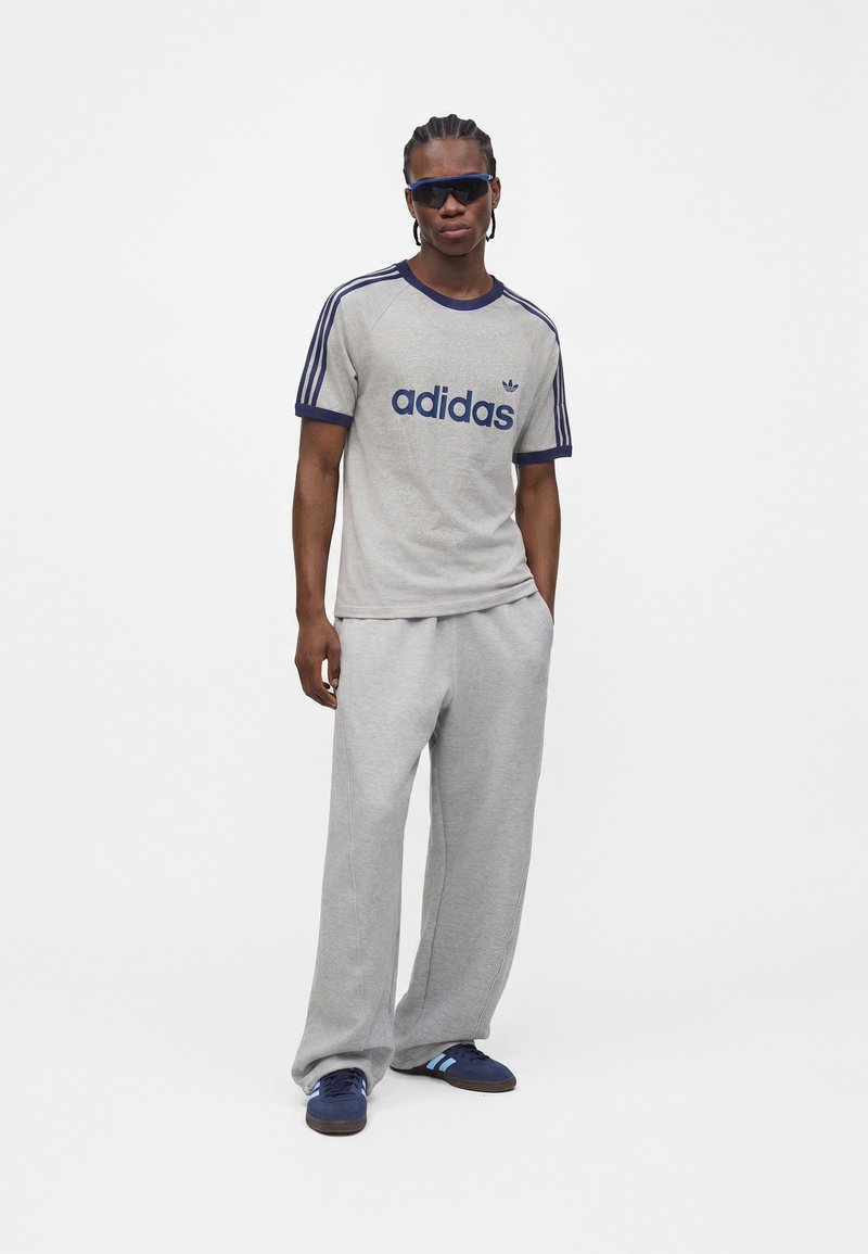 Mann trägt graues Adidas T-Shirt, hellgraue Jogginghose, dunkelblaue Sneaker und dunkle Sonnenbrille, steht vor weißem Hintergrund.