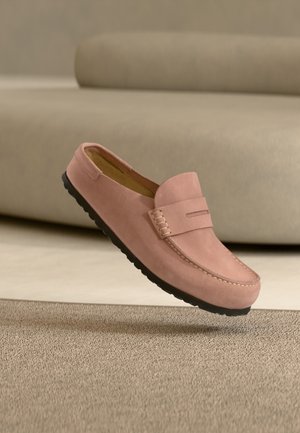 Mocasini roz din piele întoarsă cu talpă de cauciuc neagră, plutind deasupra unui covor bej, canapea bej în fundal.