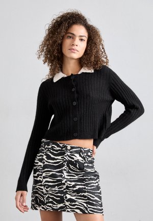 Schwarze gerippte Strickjacke mit einem cremefarbenen Kragen und sechs Knöpfen, kombiniert mit einem schwarz-weißen Minirock im Zebra-Print mit Fronttaschen.