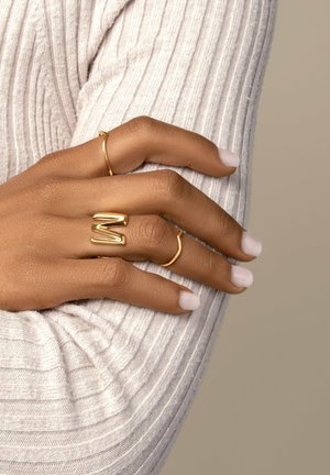 Gouden ringen op een hand: één eenvoudige dunne band en één dikker, gevormd als een 'M'. Lichtjes tekstuur op de huid en korte, lichtgekleurde nagels.
