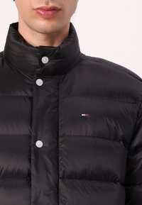 Svart dunjakke med høy krage, trykkknapper, quiltet design og et lite logopatch på venstre bryst. Jevn tekstur.