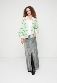 Πλεκτή ζακέτα σε ανοιχτό κρεμ με πράσινες ρίγες, συνδυασμένη με μακριά, υφασμάτινη φούστα με γκρι σχέδιο φιδιού και μαύρες μπότες μέχρι το γόνατο.