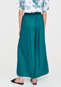 Szerokie spodnie w odcieniu zielono-niebieskim z marszczonym elastycznym paskiem, wykonane z gładkiego materiału. Noszone z czarnymi klapkami, widok od tyłu.