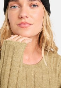 Olivgrüner, gerippter Pullover mit langen Ärmeln, zeigt eine weiche Textur. Die Hand des Modells ruht auf dem Kinn, während es eine schwarze Mütze trägt. Neutraler Hintergrund.