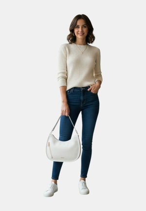 Donna con maglione beige a coste e jeans skinny blu scuro che tiene una borsa bianca con manici grigi, indossa sneakers bianche e gioielli dorati.