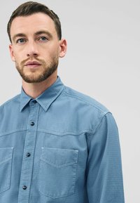 Een man met een korte baard en net haar draagt een blauwe overhemd met een gestructureerd patroon. Hij kijkt rechtstreeks naar de kijker tegen een effen achtergrond.