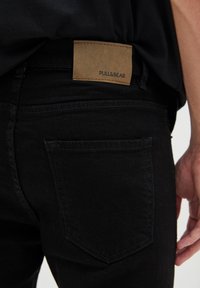 Jeans negros con un acabado texturizado, que presentan un parche de cuero marrón con el logo "PULL&BEAR" en relieve, realzado por bolsillos traseros visibles.