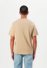 Beige pamut póló rövid ujjakkal és kerek nyakkivágással, hátulról bemutatva, világoskék farmernadrággal párosítva.