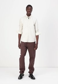 Chemise à rayures couleur crème clair, assortie à un pantalon marron. Des chaussures noires complètent la tenue. Le mannequin se tient debout, les bras détendus le long du corps.