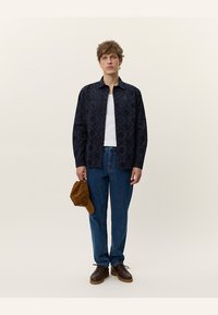 Marineblå skjorte med knapper og mønstrete tekstur, hvit tanktop, blå jeans, brune sko og en beige caps holdt i hånden. Enkle farger og uformell stil.