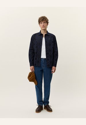 Marine blouse met een geblokt patroon, witte tanktop, blauwe jeans, bruine schoenen en een tan pet in de hand. Effen kleuren en casual stijl.