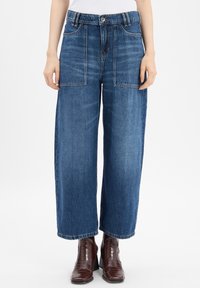 Wijd-uitlopende, mid-blauwe denim jeans met twee grote voorzakken, riemlussen en een ruwe zoom. Gecombineerd met donkerbruine enkellaarsjes.