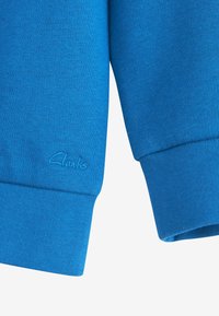 Sinised pusa varrukad soonikuga mansettide ja väikese tikkitud Clarks logo vasakul mansetil.