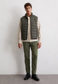 Grüne gesteppte Weste über einem beigen Langarmshirt, kombiniert mit grüner Hose und schwarzen Schuhen. Einfaches Design und moderner Schnitt.