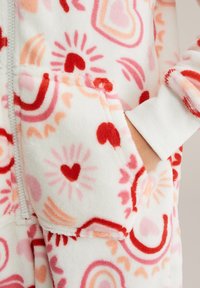 Mono de felpa blanco con corazones multicolores y patrones abstractos. Cuenta con una cremallera frontal y puños elásticos. Tela suave y texturizada.