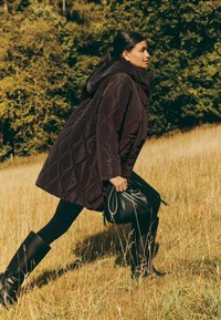 Prešívaný tmavofialový vatovaný kabát s vysokým golierom a kapucňou; v kombinácii s čiernymi legínami a čiernymi čižmami po kolená; drží čiernu tašku.