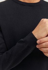 Czarny dziany sweter z okrągłym dekoltem i ściągaczami na mankietach. Gładka faktura i dopasowane rękawy. Podkreślony detal na nadgarstku pokazuje czyste szwy.